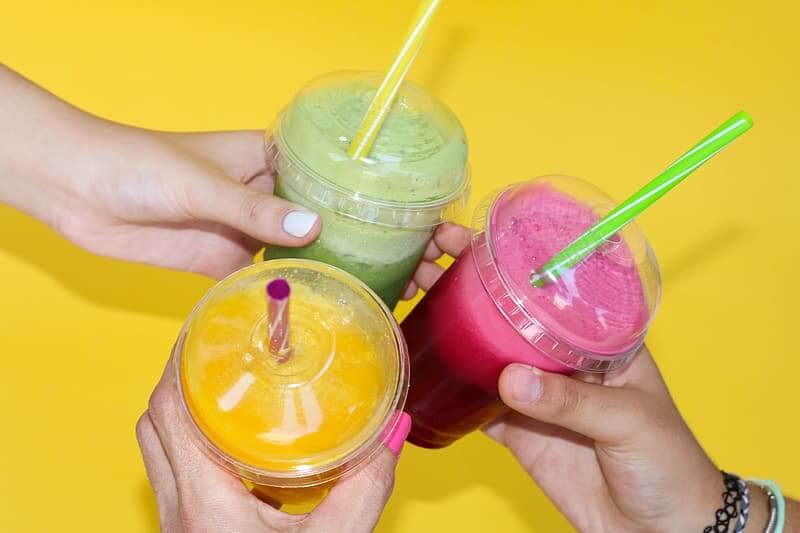 Tiga tangan memegang tiga jenis smoothie hijau, merah, dan kuning